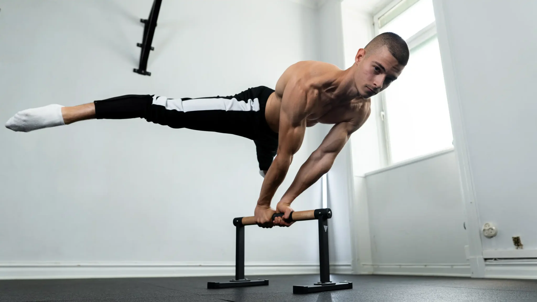 Daniel Hristov holding a straddle planche on parallettes in a bright white room