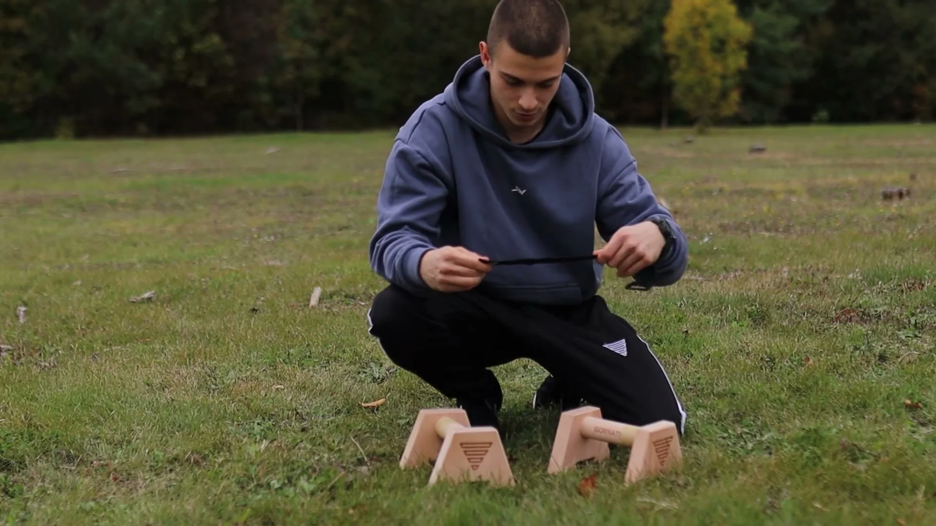 Daniel Hristov assembling a pair of wooden GorNation parallettes on grass, fitting the crossbar into the triangular base